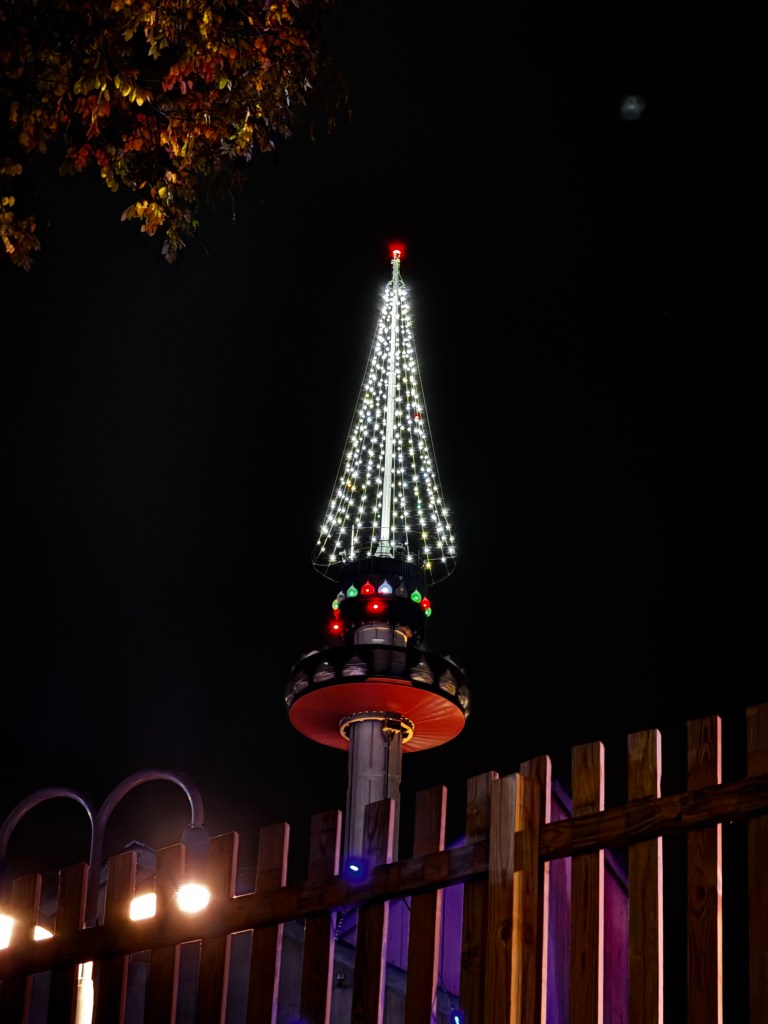 A brightly lit Kissing Tower observation ride at night, featuring a conical design adorned with colorful lights, set against a dark sky.