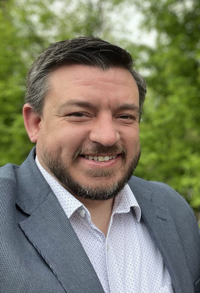 A man with short, neatly styled hair and a beard smiles for the camera, wearing a light-colored dress shirt and a blazer, with green foliage in the background.